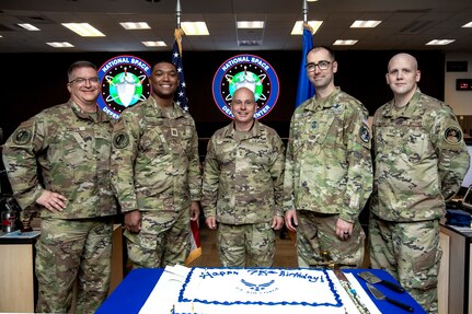 Five men in military uniforms pose for a photo standing in front of a table with a sheet cake