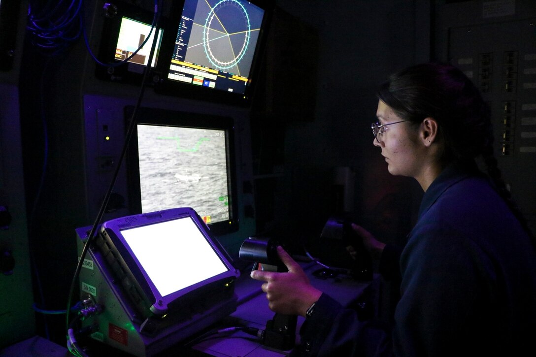 A sailor operates a weapons system with screens inside a dark room.