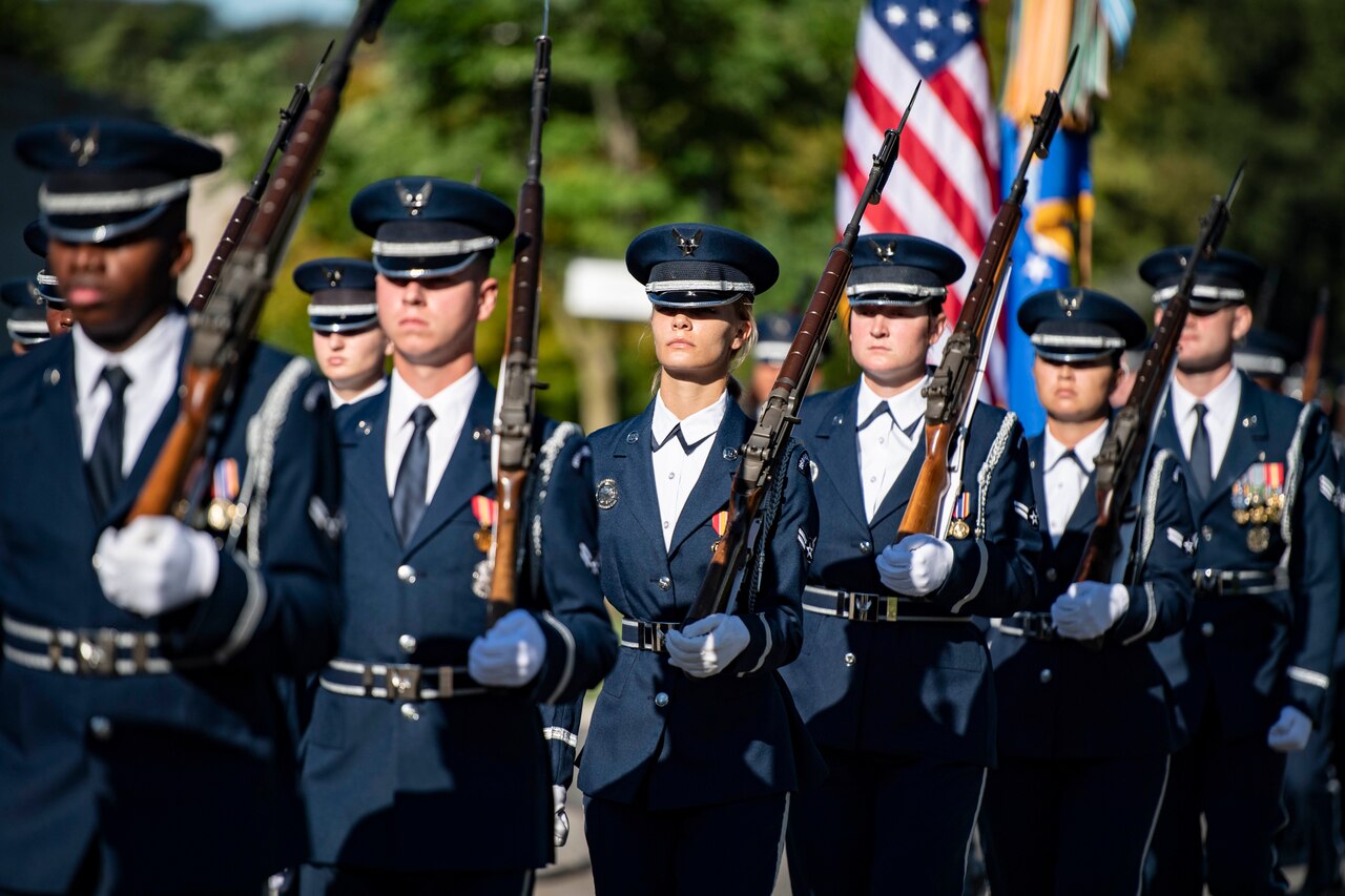 A group of airmen walk in formation carrying rifles and flags.