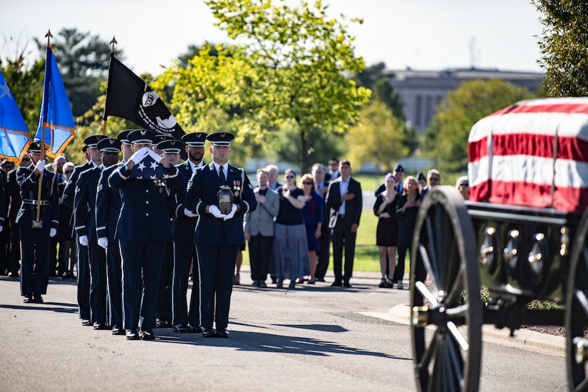 Service members walk behind a flag-draped caisson, some with flags while people stand on the sidelines.