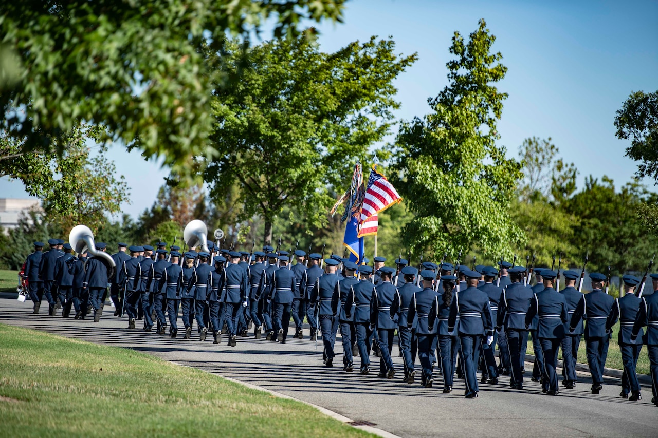 A procession of airmen depart a cemetery carrying a flag and musical instruments.
