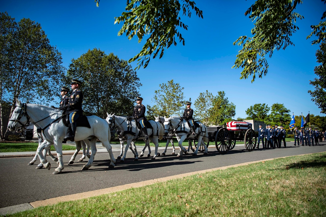 Service members on horses pull a flag-draped casket followed by others on foot with flags.
