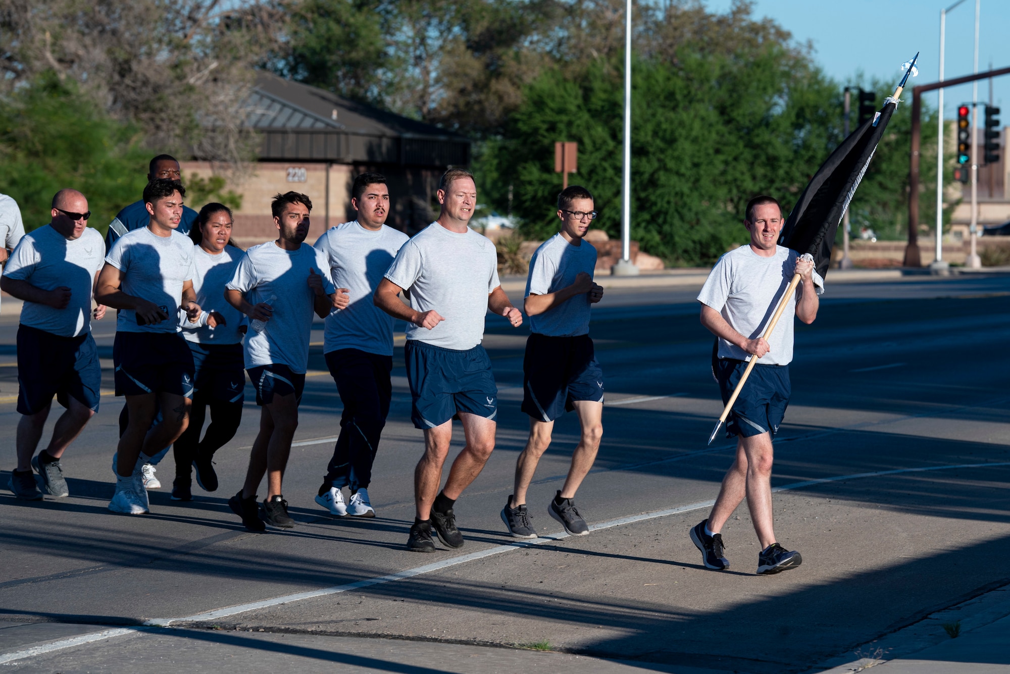 A group of people in grey Air Force physical training uniforms run with the leading person carrying a black and white POW/MIA flag.