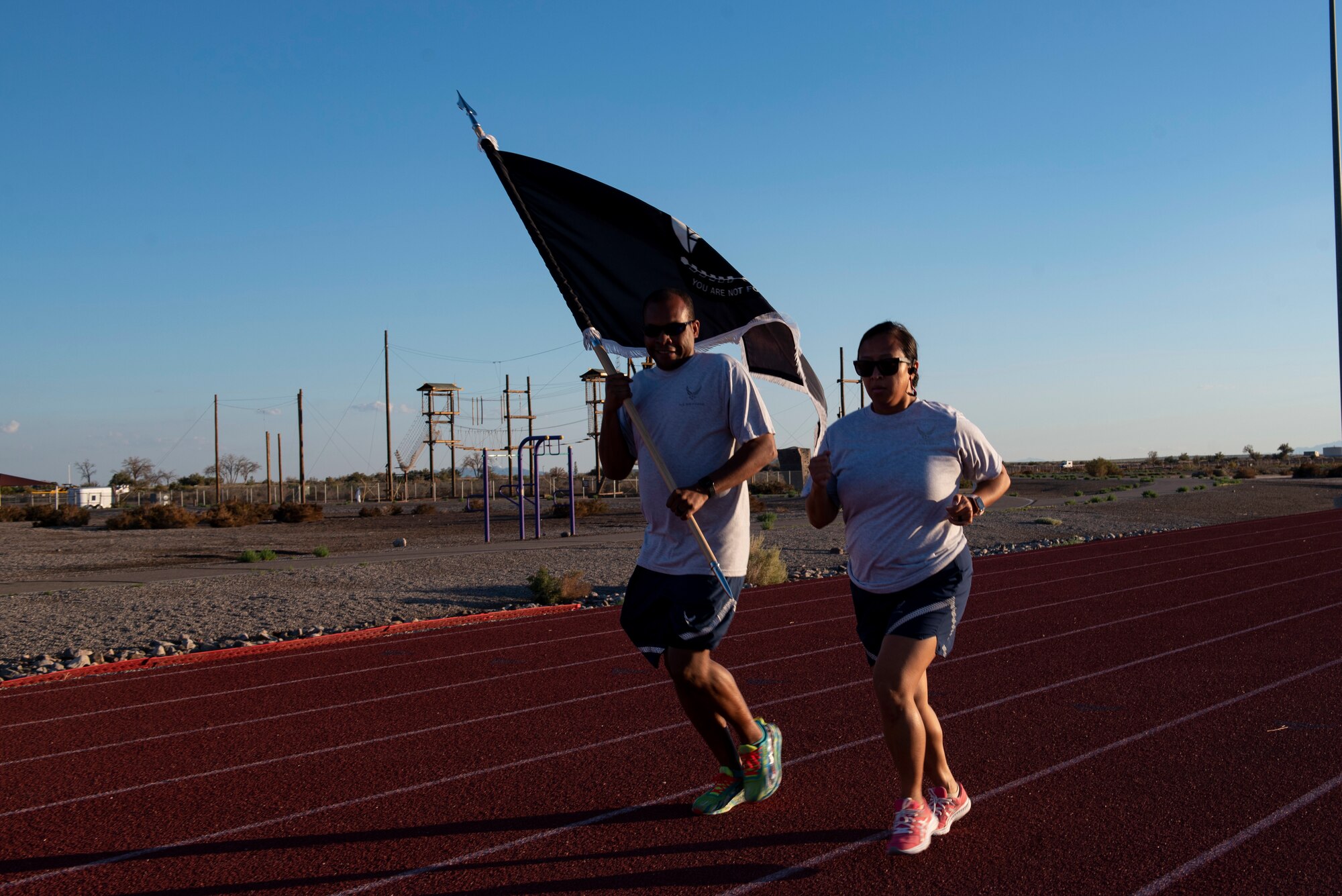 Two people in grey and blue Air Force physical training gear run on a red track with a black and white POW/MIA flat.