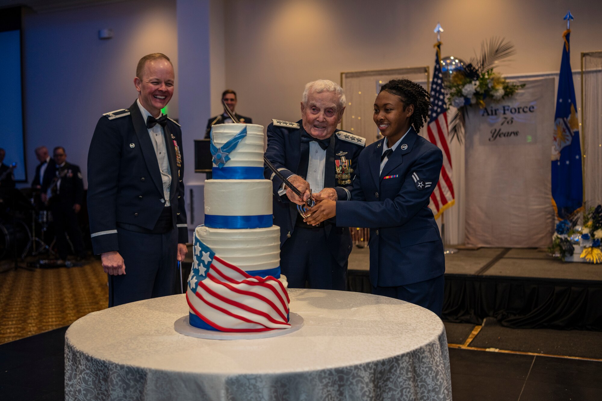 Two men and a woman stand around a cake as they cut the cake with a large military style sword.