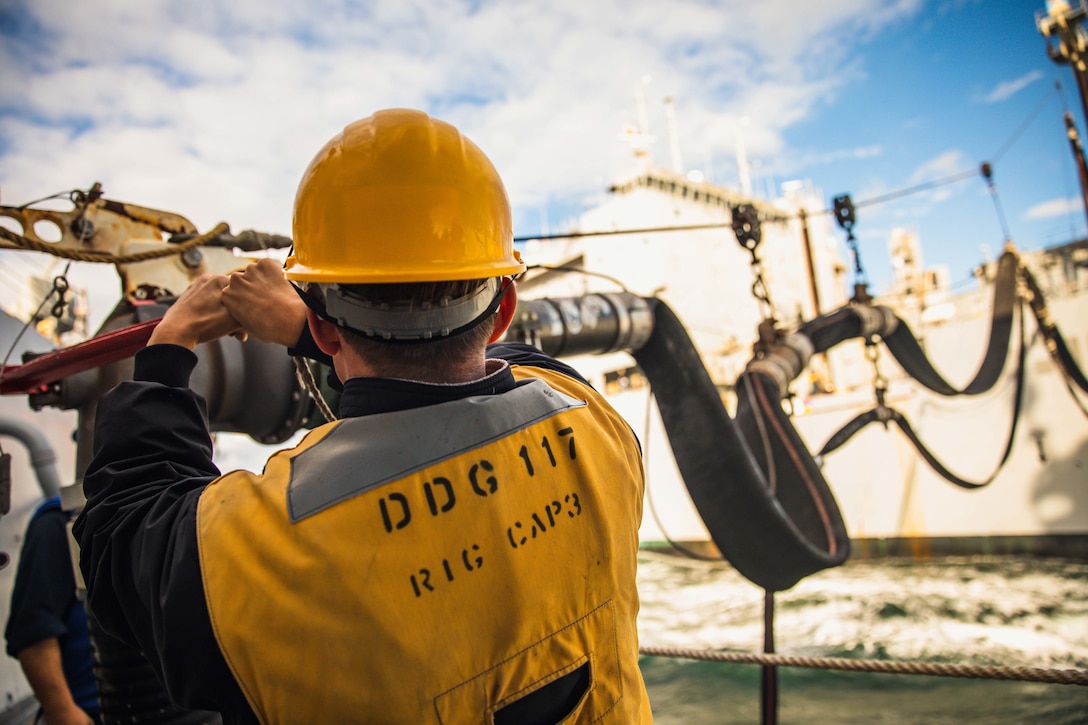 A sailor tends to a fuel line coming from an adjacent ship at sea.