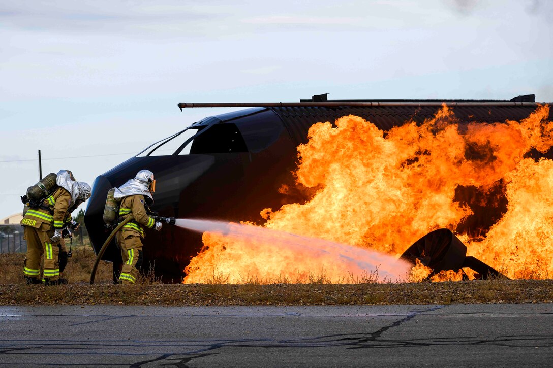 An airman wearing fire protection gear uses a fire hose on an aircraft fire as two others stand behind.