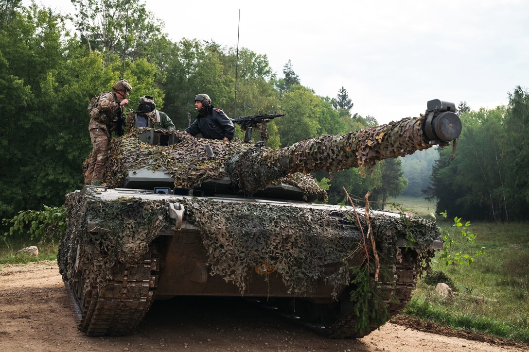 A soldier stands on the body of a tank and talks to the two soldiers operating the vehicle.