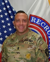 man wearing u.s. army uniform standing in front of two flags.