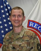 man wearing u.s. army uniform standing in front of two flags.