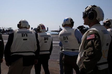 Three men watch an aircraft on the deck of an aircraft carrier.
