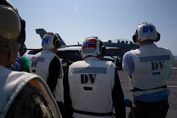 Three men watch and aircraft on the flight deck of a ship.