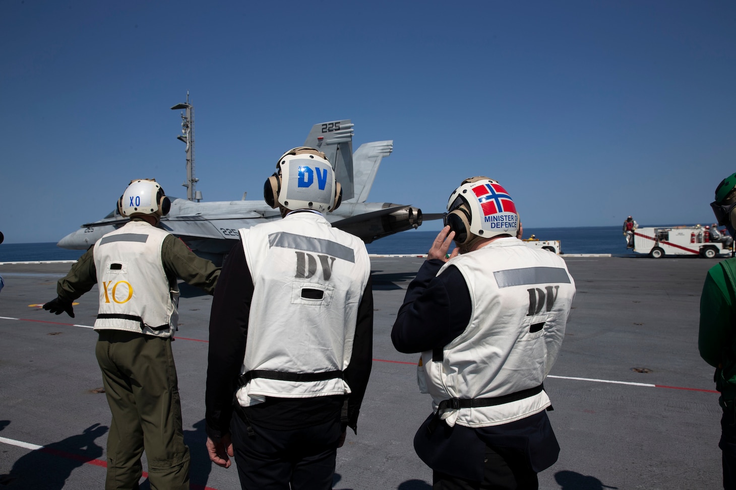 Three men watch an aircraft on the deck of an aircraft carrier.