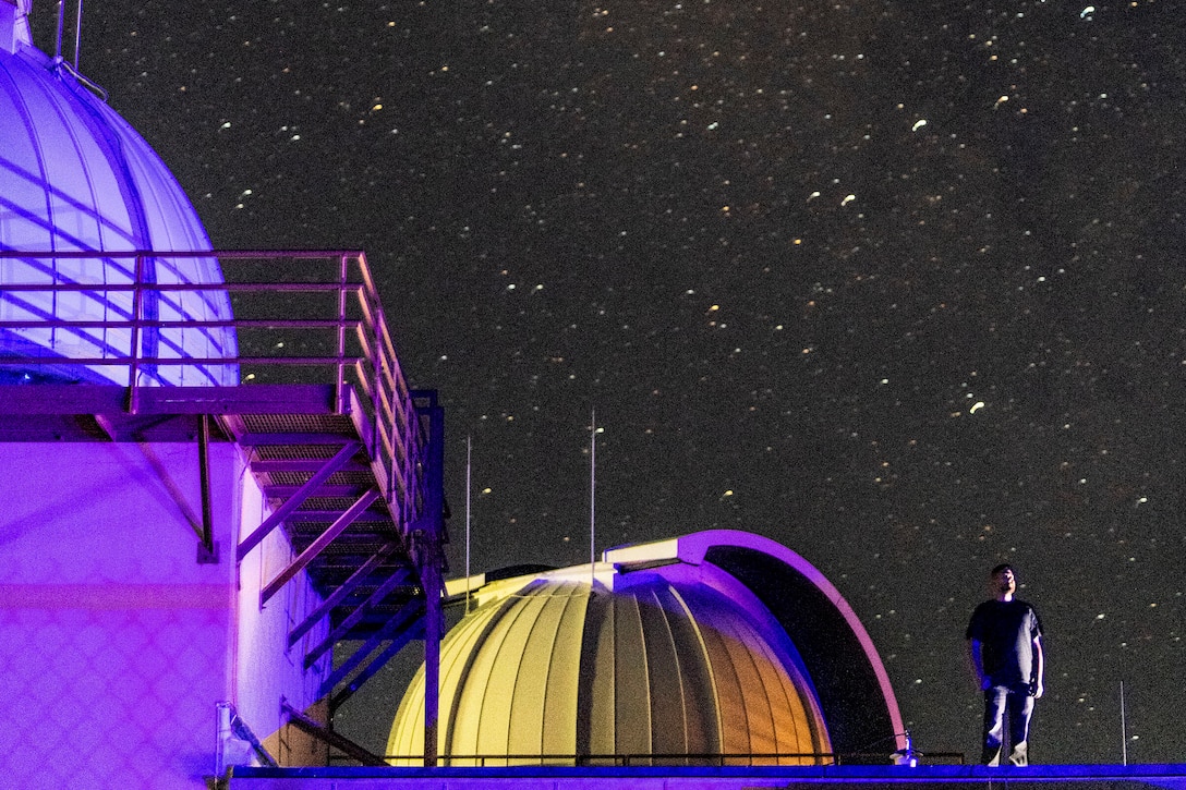 A person looks at a starry night sky while standing by a domed building illuminated by purple light.