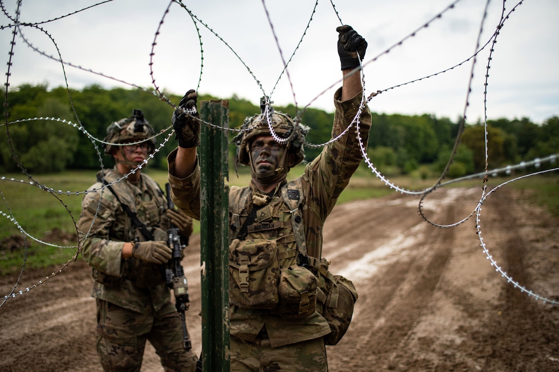 One soldier holds and wraps concertina wire while another soldier looks on.