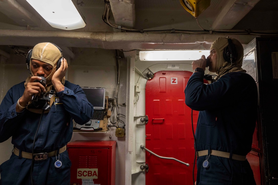 Two sailors wearing navy blue coveralls and tan flash hoods talk on communication devices in a ship.