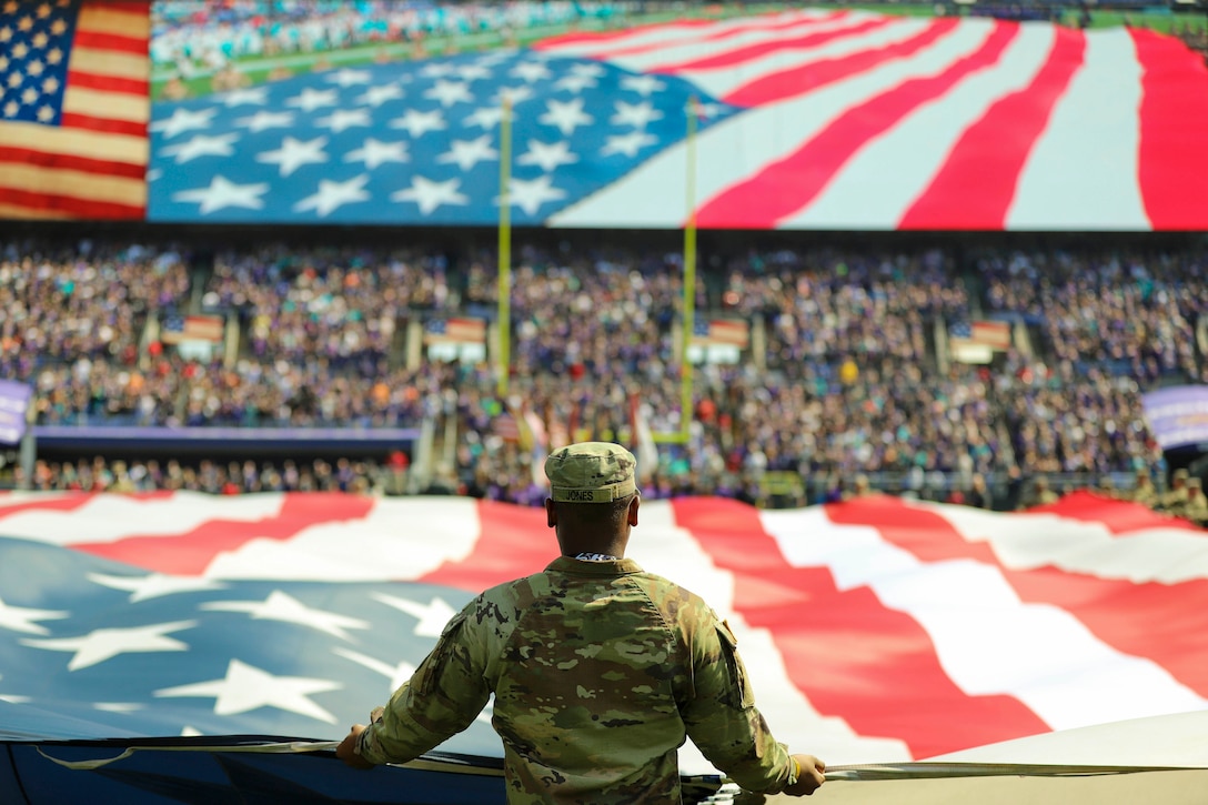 A National Guardsman holds a large American flag on a football field as fans sit in the stadium.