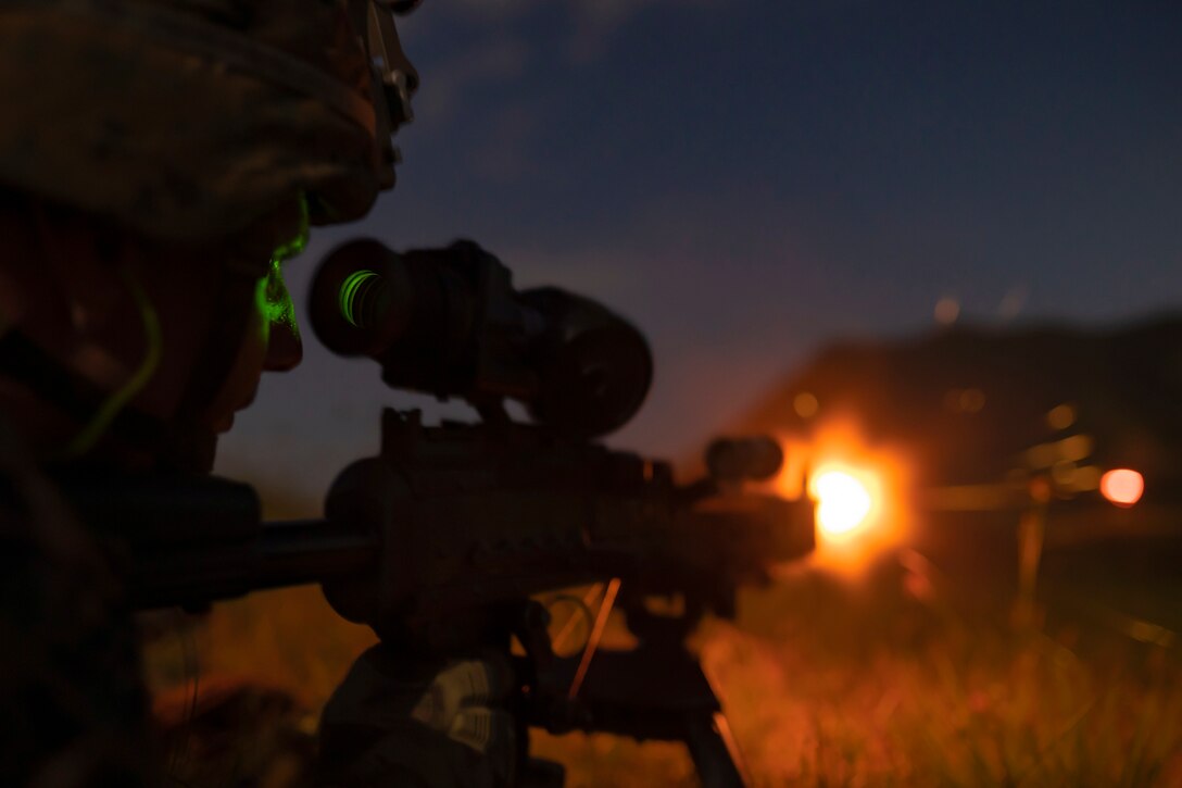 A Marine looks through a viewfinder before firing a weapon in a field in the dark.