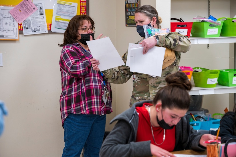 A service member holding paperwork talks to a woman in a classroom.