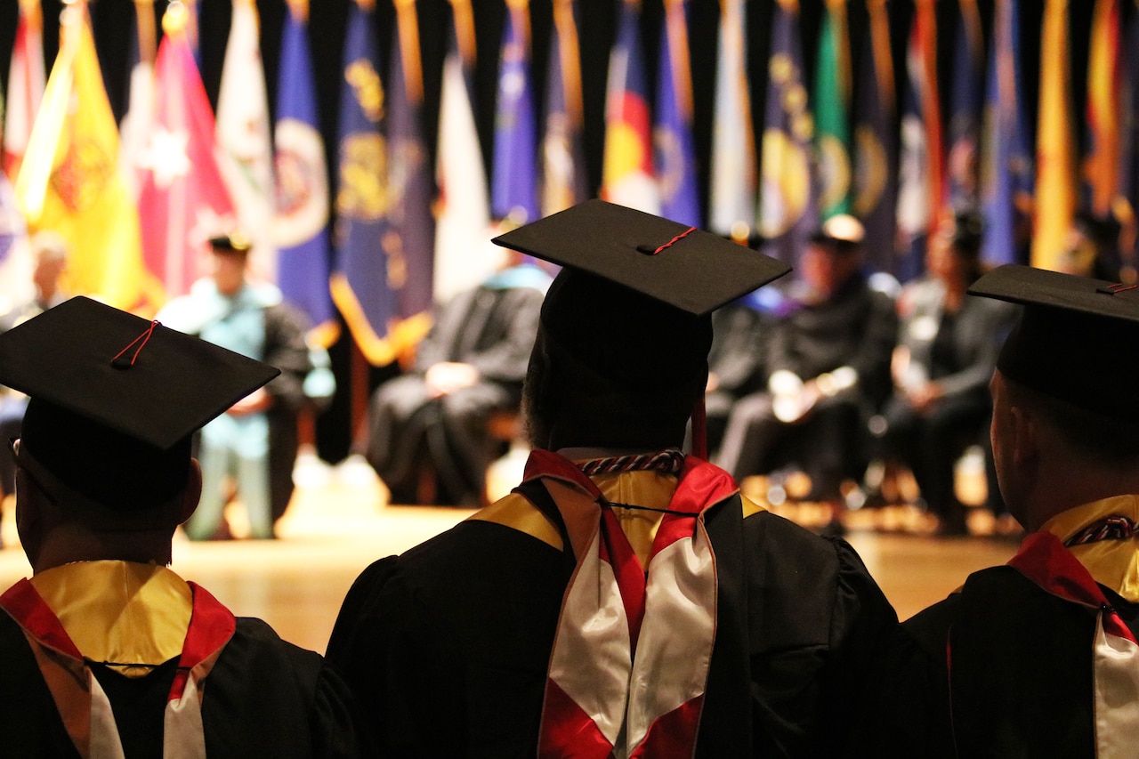 People wearing caps and gowns look toward a people seated on stage adorned with flags.