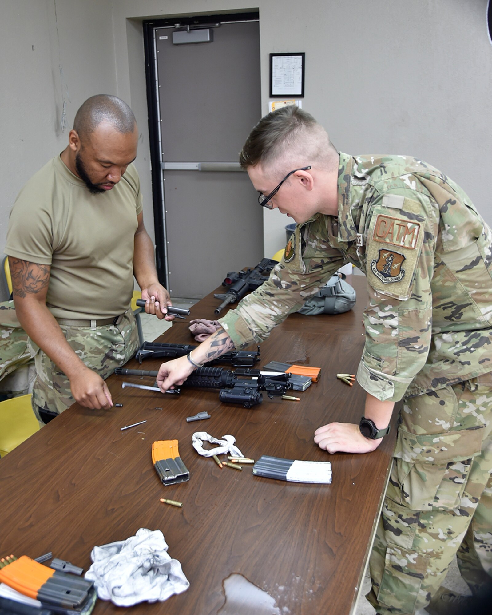 Two male servicemembers stand on either side of a table, man on left holds bolt assembly of M-4 rifle while man on right instructs on what piece to use next.