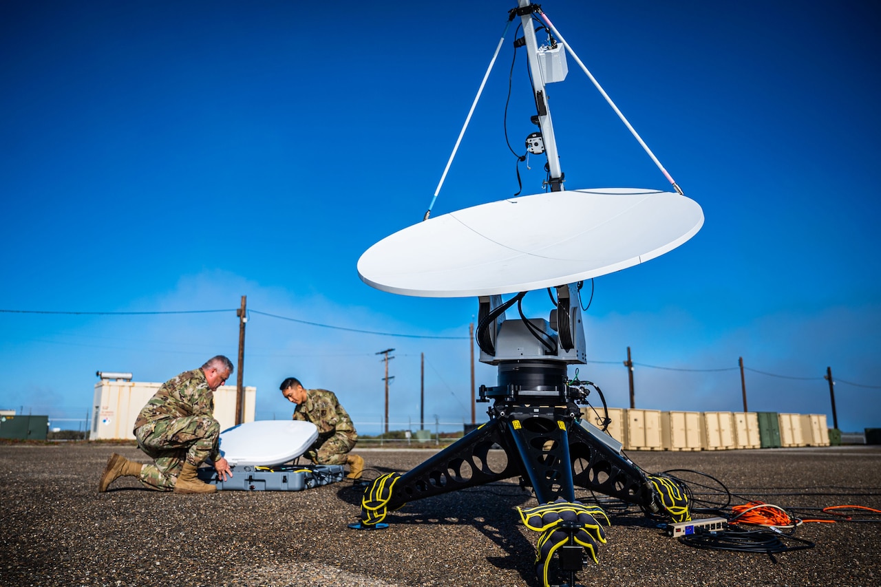 Two service members handle electronic gear on the ground near a portable antenna.
