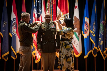 man wearing u.s. army uniform standing on a stage while two people pin a star on each shoulder.