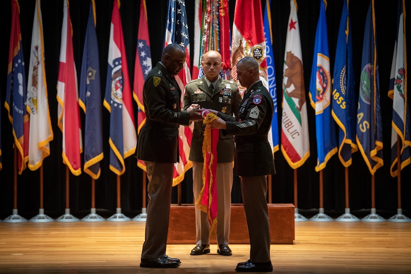man wearing u.s. army uniform standing on a stage unrolls a flag with another man wearing an u.s. army uniform.