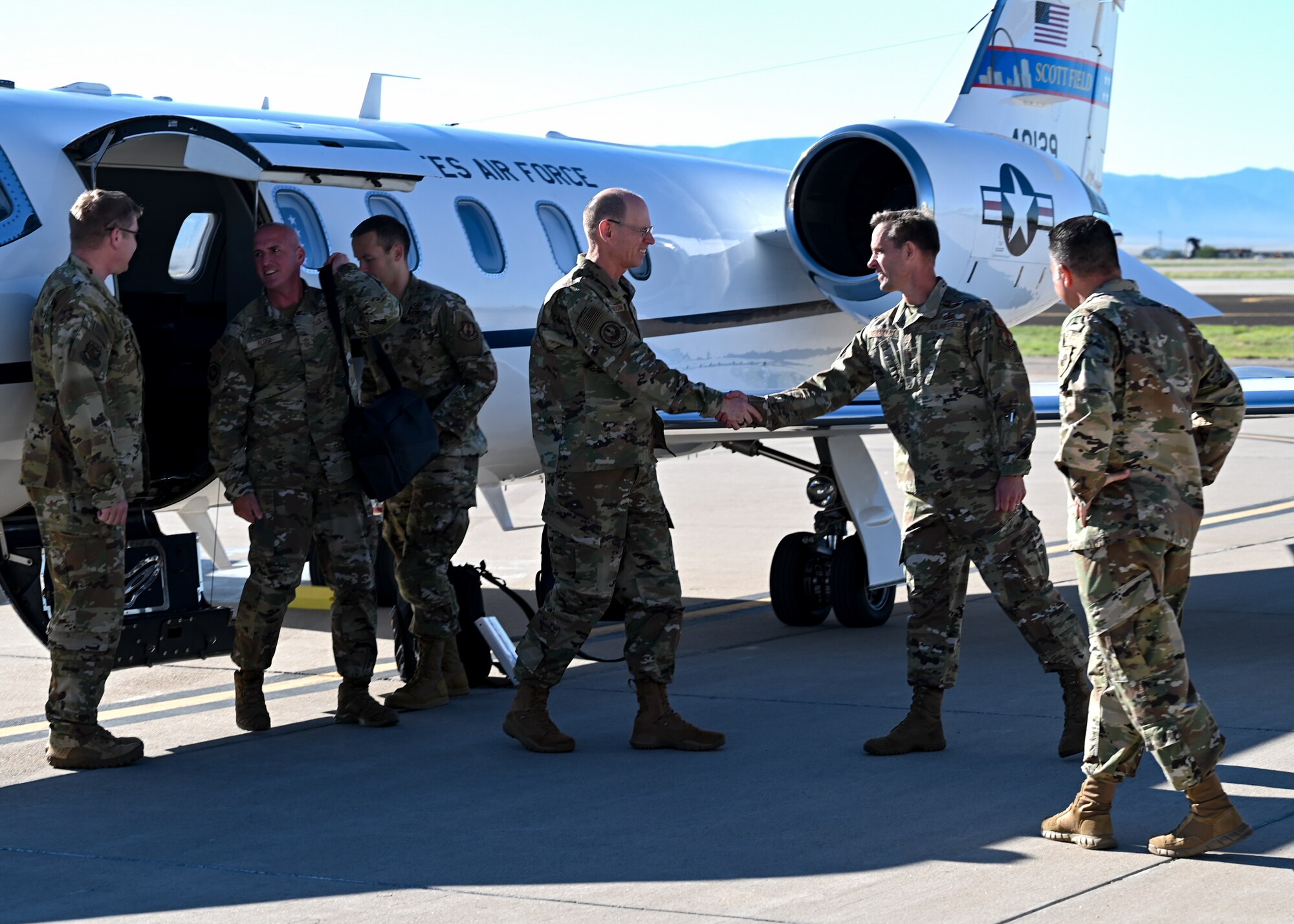 Two Airmen in uniform shake hands in front of a plane. One more is leaving the plane while the other three wait to greet the arrivals.