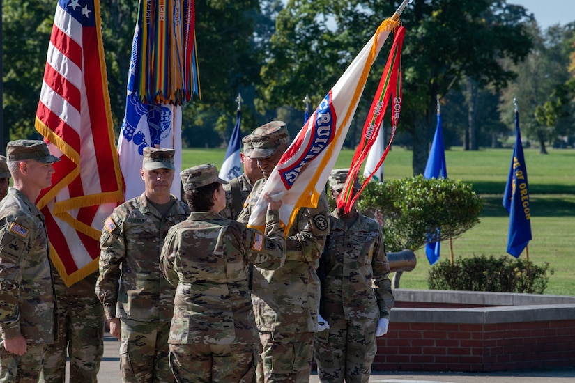 man in u.s. army uniform holding flag passes it to woman in uniform.