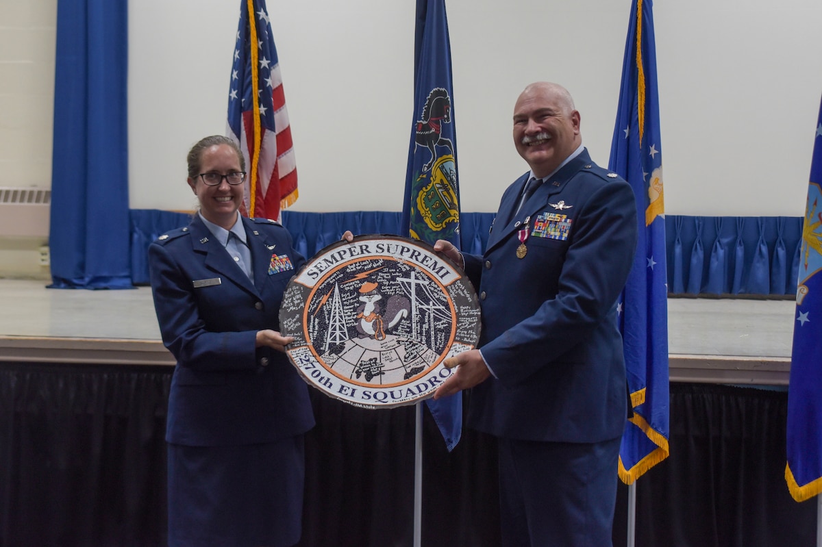 A woman in Air Force dress blues hands a unit logo to a man in Air Force dress blues.
