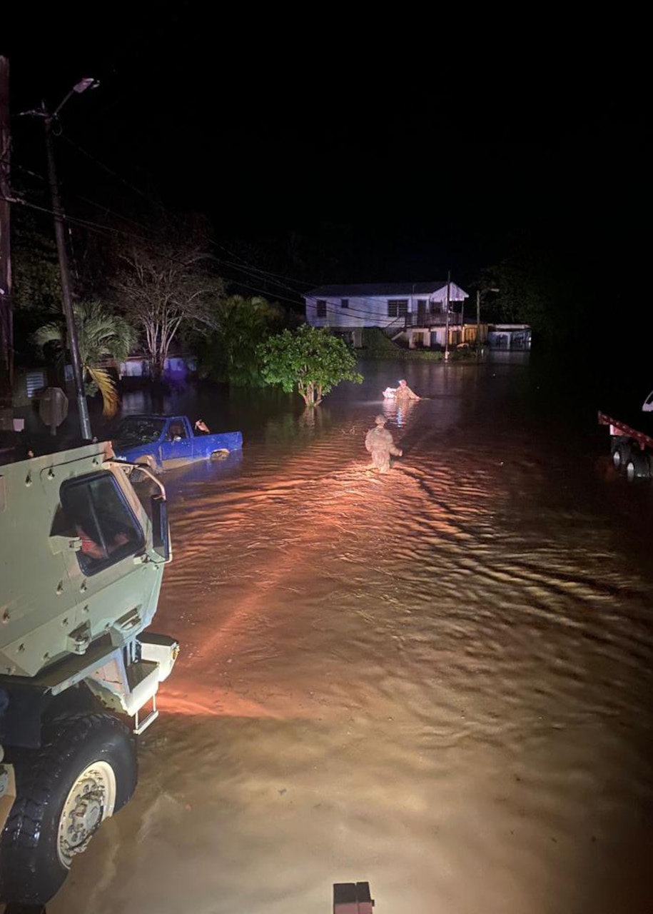 Service member approaches person in knee-deep water on street as military vehicle sits nearby in dark.