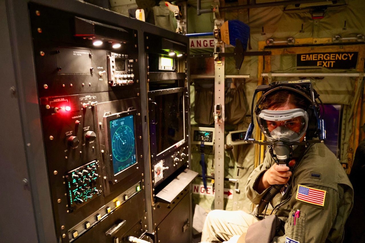 An airman seated by an aircraft control panel wears an oxygen mask