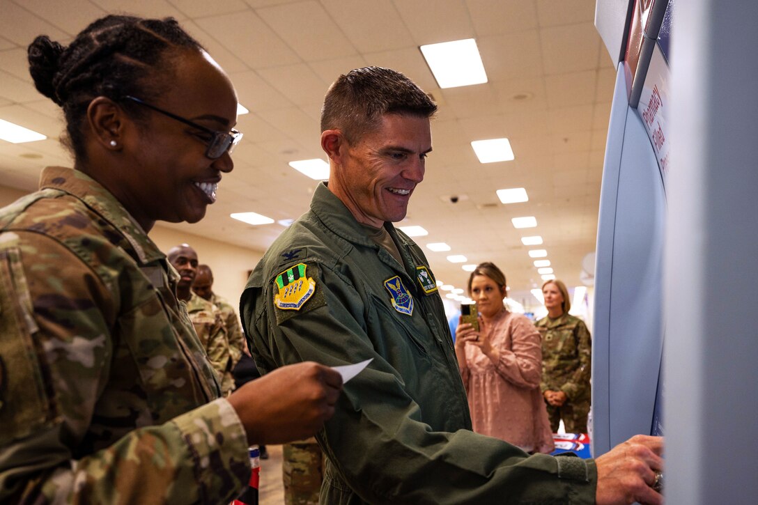 Two smiling service members stand at an indoor kiosk as other people watch.
