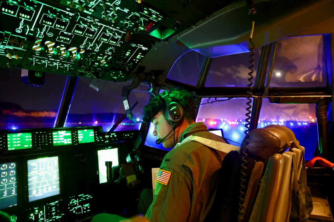 An airman reviews a control panel in a cockpit illuminated by green lights.