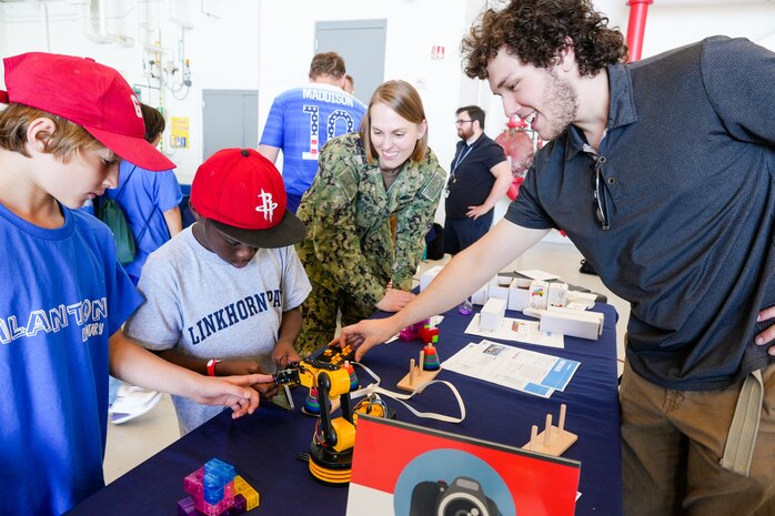 IMAGE: Naval Surface Warfare Center Dahlgren Division Dam Neck Activity Commanding Officer Cmdr. Christina Carino and intern Johnathan Gilliam guide a Linkhorn Park Elementary School fifth-grader as he uses a robotic claw to move various shaped blocks from one post to another during the first day of the Naval Air Station Oceana Air Show.