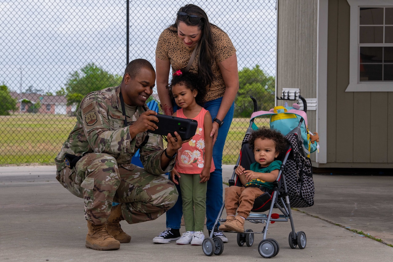 A uniformed service member kneels while a small child and woman look at the device in his hand.