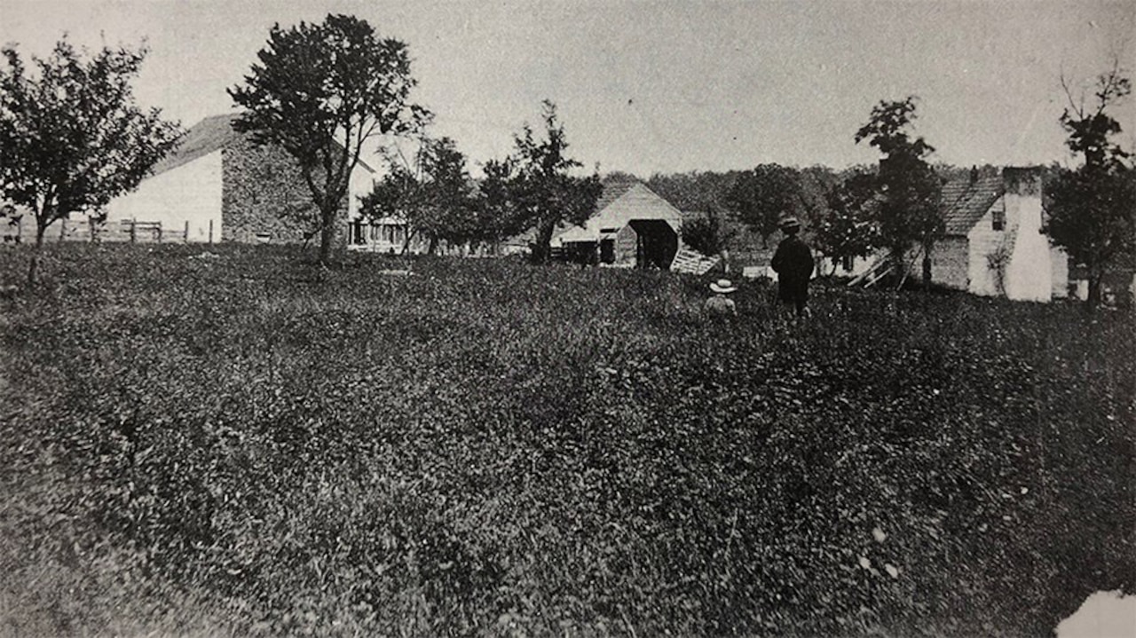 Two people walk through a field with barns in the background in a black and white photo.