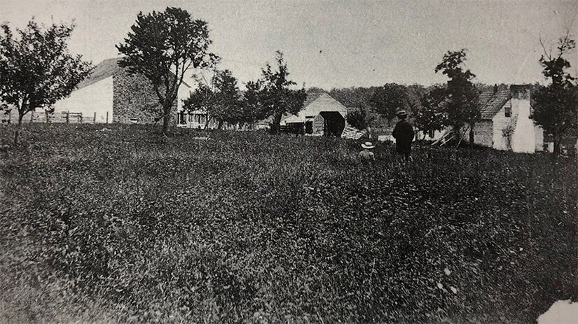 Two people walk through a field with barns in the background in a black and white photo.