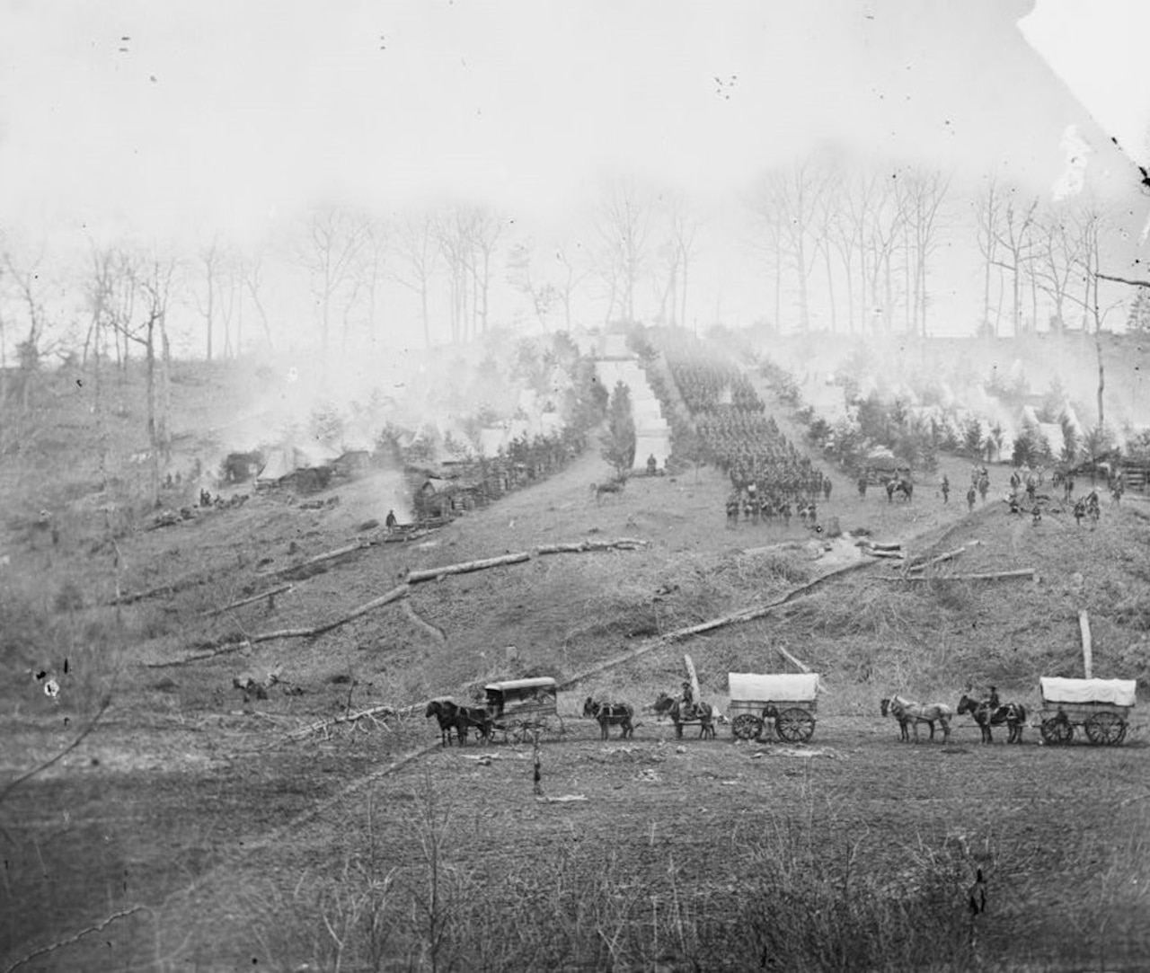 A campsite sits on a hill with covered wagons and horses in the foreground in a black and white photo.