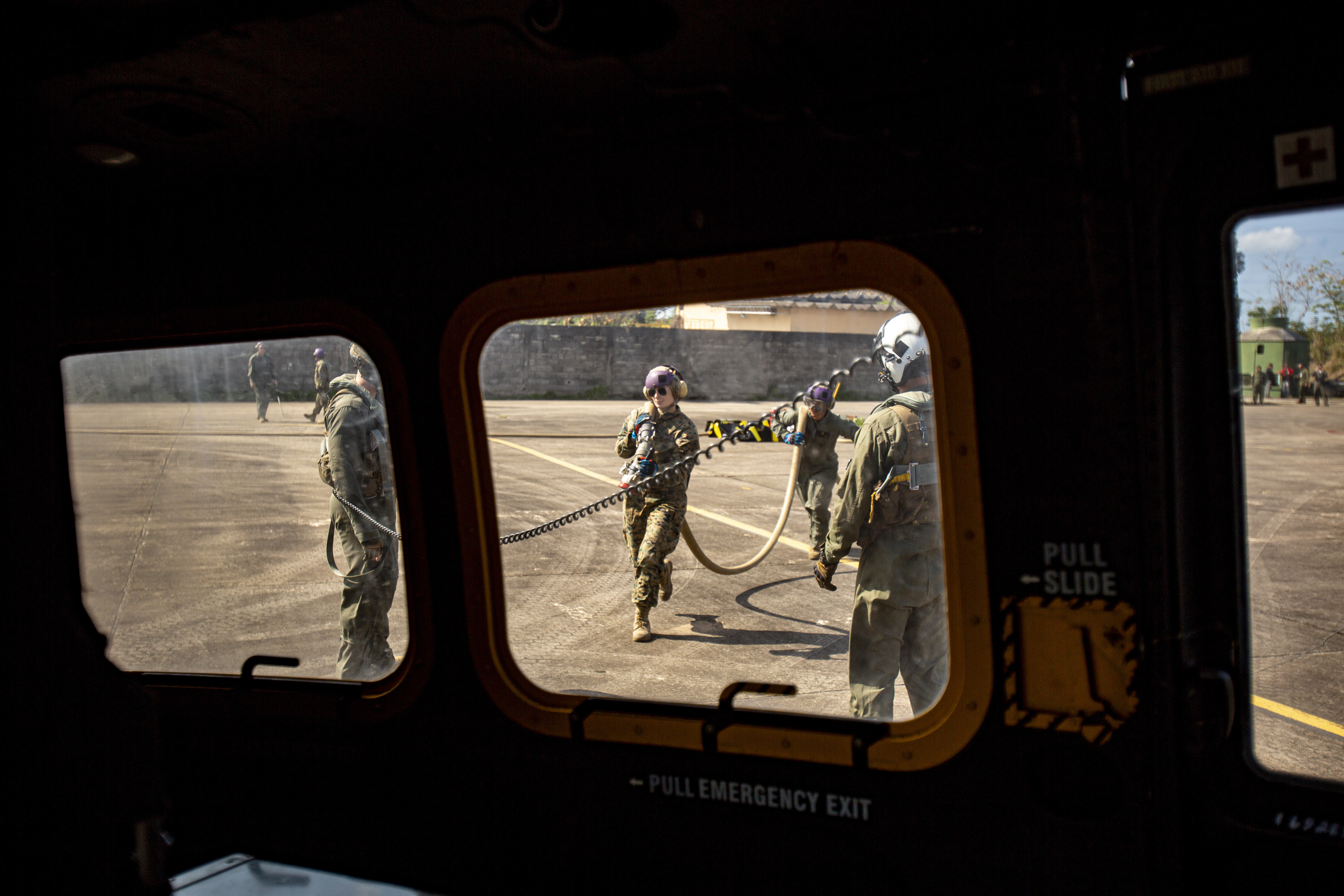 Marines holding a fuel line outside, from the point of view of inside an aircraft through windows.