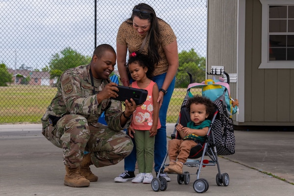 A uniformed service member kneels while a small child and woman look at the device in his hand.