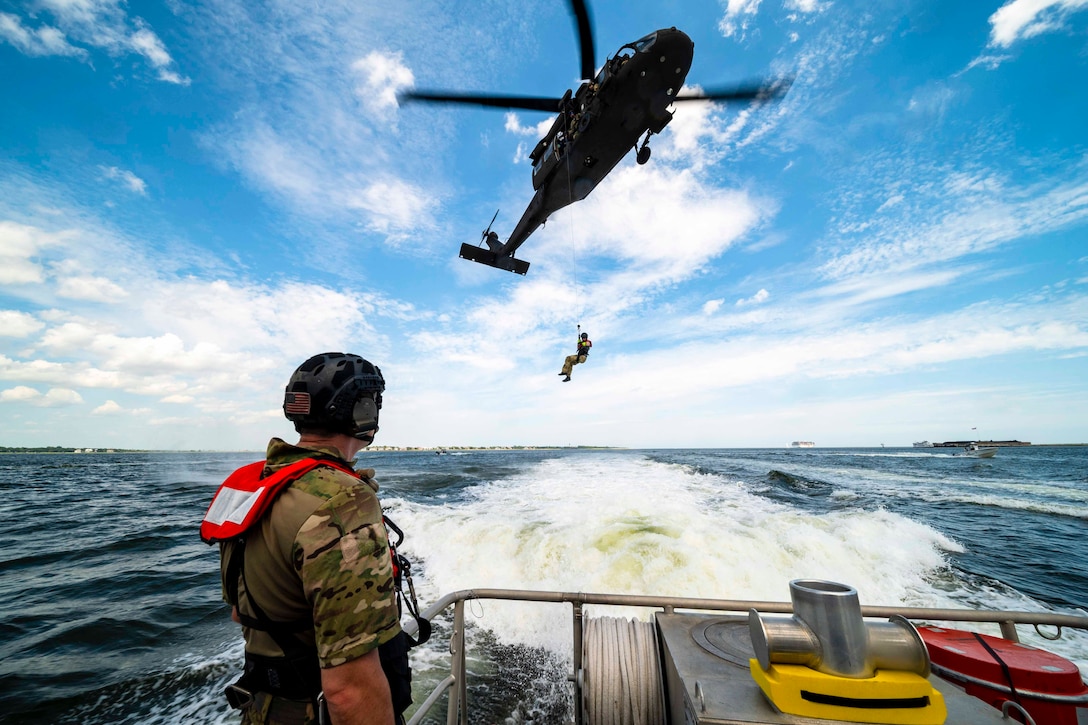 A person is lowered from an airborne helicopter to a ship where another person watches from the deck.