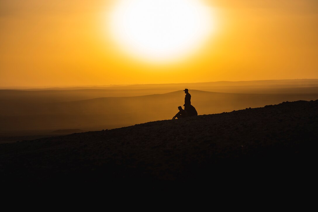 Two soldiers sit and one soldier stands while watching a sunrise.