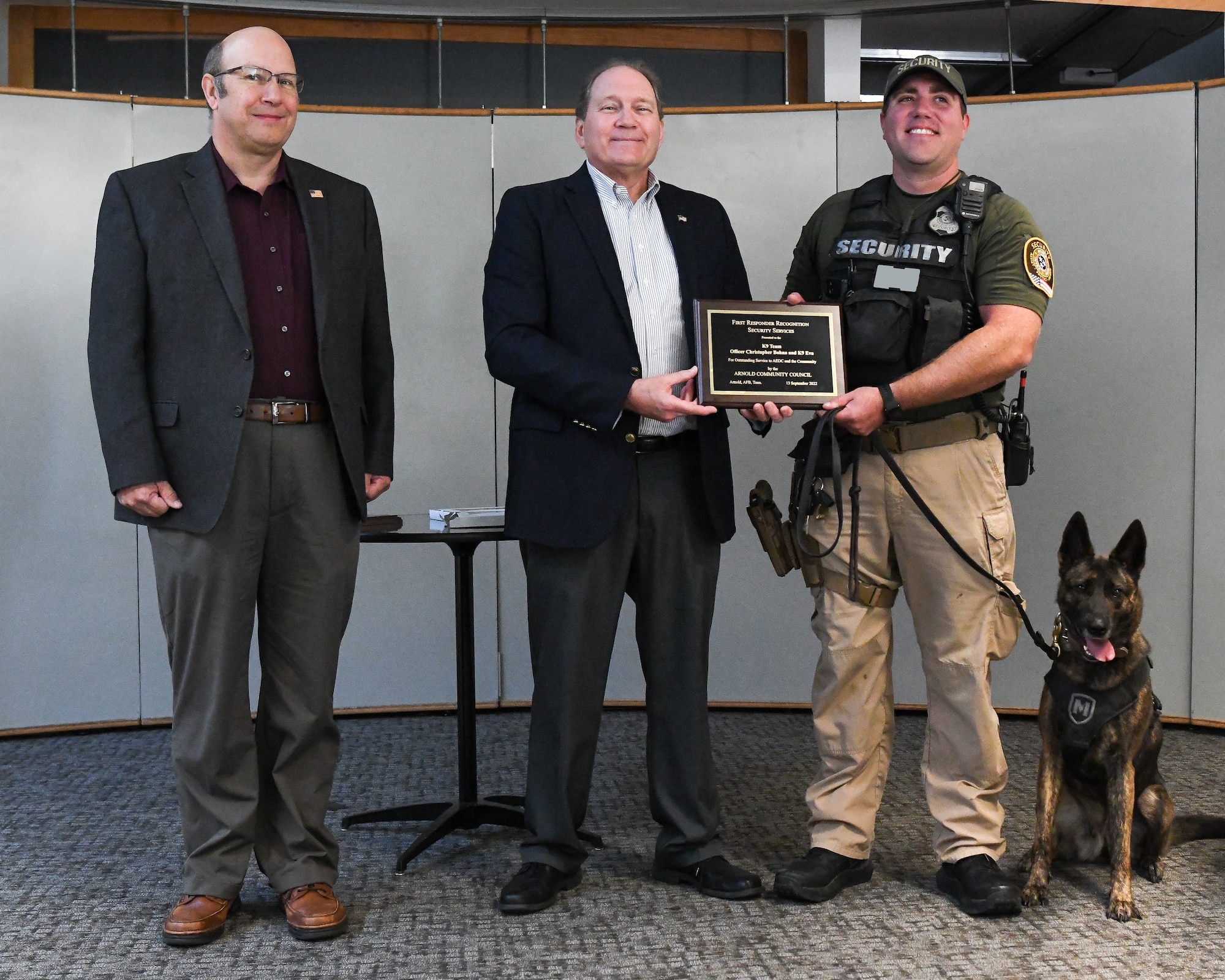 Man presents plaque to security guard and K-9, with another man pictured.