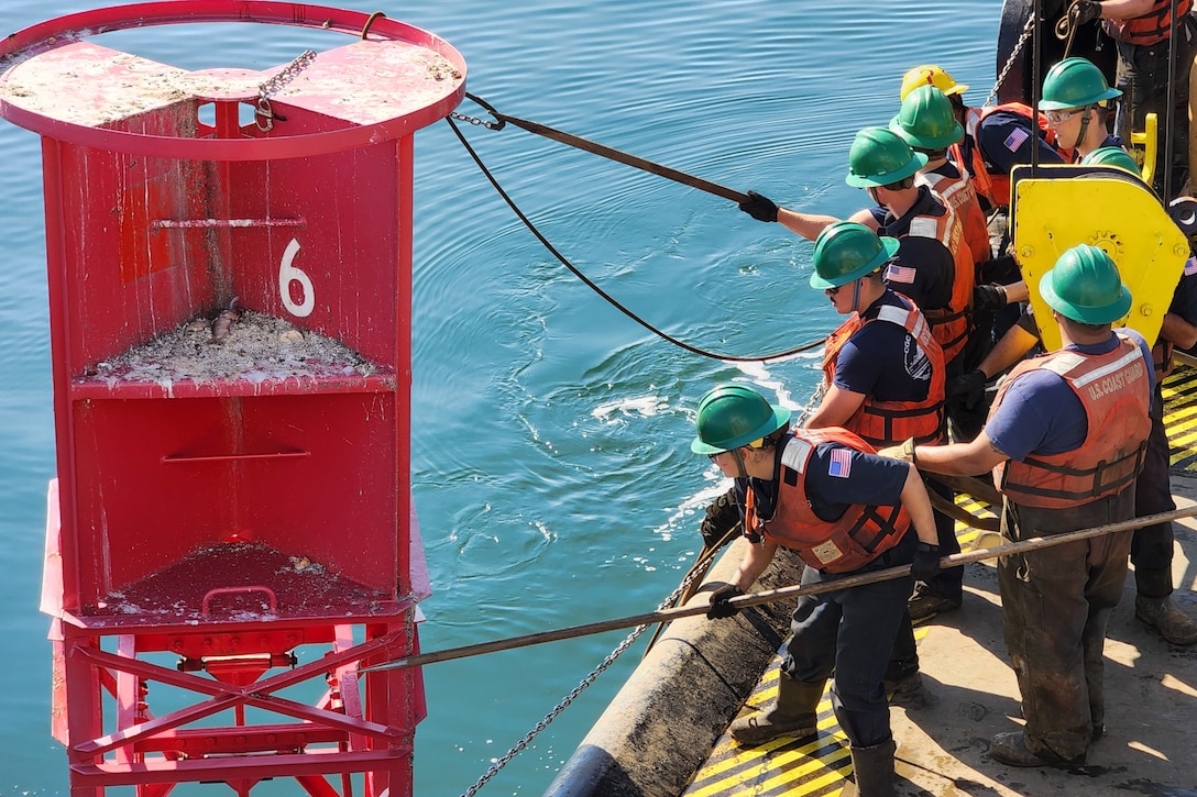 Members of the Coast Guard use hooks and rods to being a large buoy on board a ship.