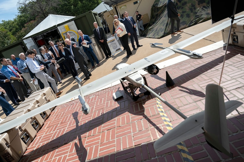 A large group looks at a drone sitting on the ground.