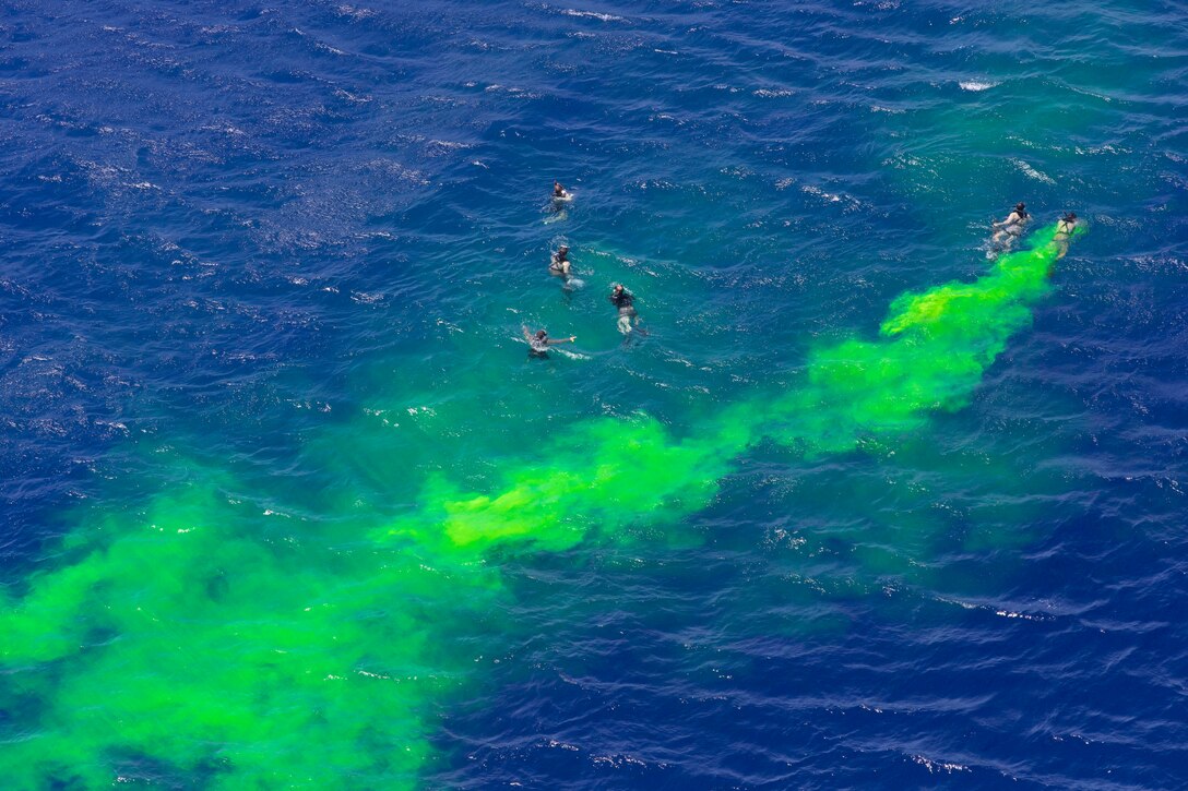 Overhead view of swimmers in vivid blue water with bright green smoke billowing above them.