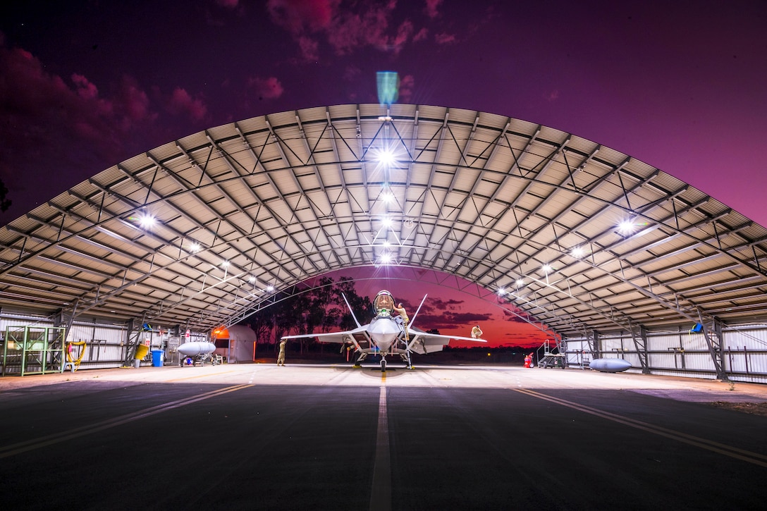 Airmen inspect an aircraft under a curved roof against a purple sky.