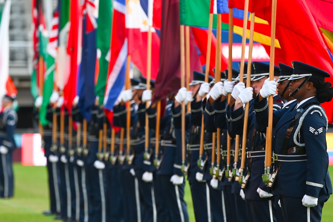 Airmen stand in a line on a field and hold up national flags.